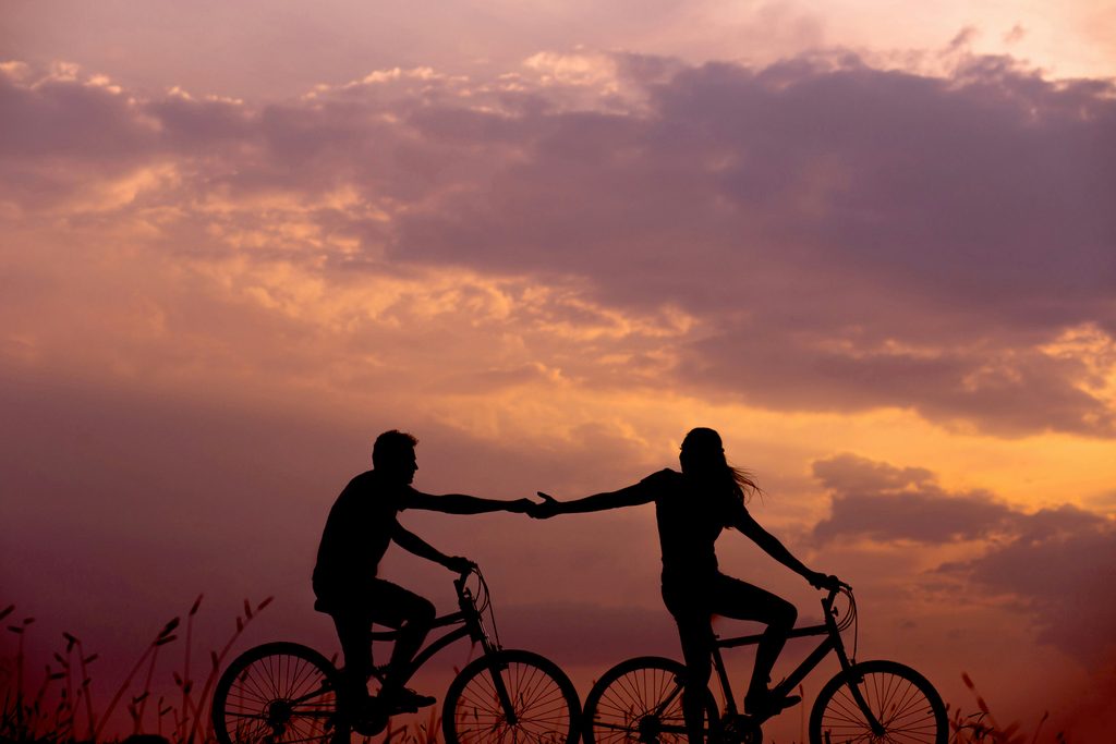 Silhouettes of a man and woman riding bicycles together at sunset, reaching toward each other as they cycle beneath a dramatic sky filled with warm orange, pink, and purple clouds.