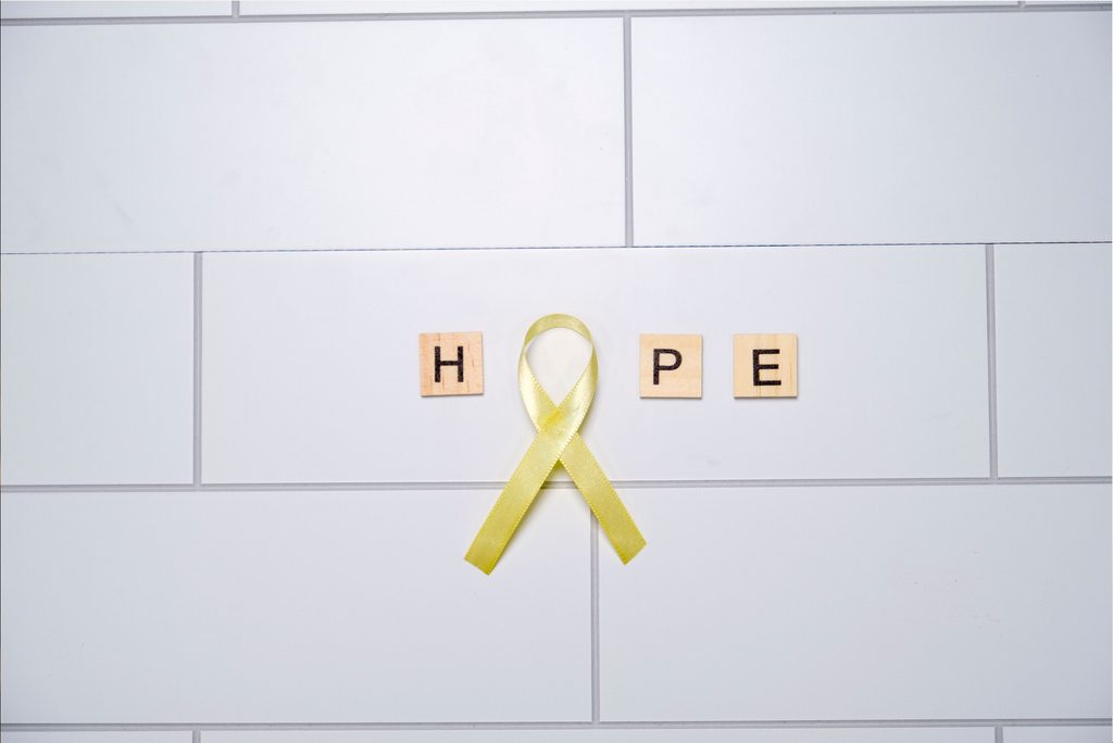 A yellow awareness ribbon placed between wooden letter tiles spelling “HOPE” on a white tiled background, symbolizing hope, support, and awareness.