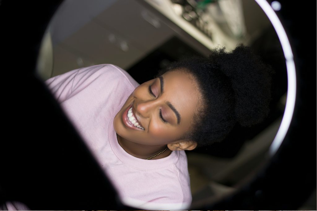 A smiling Black woman with natural curly hair wearing a light pink top, photographed through a circular ring light with her eyes closed and a joyful expression.