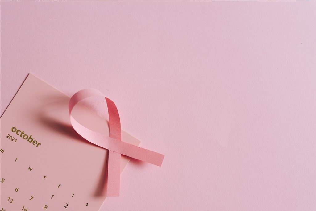 A pink breast cancer awareness ribbon rests on an October calendar against a soft pink background, symbolising breast cancer awareness and the importance of early detection.
