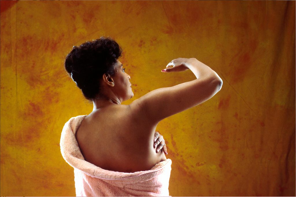A Black woman wrapped in a soft towel examines her breast while raising one arm, illustrating breast self-awareness and the importance of checking for unusual changes as part of breast cancer awareness.