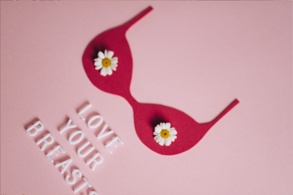 A pair of burgundy-red sunglasses decorated with white daisies rests on a soft pink background beside white lettering that reads “Love Your Breasts,” promoting breast cancer awareness.