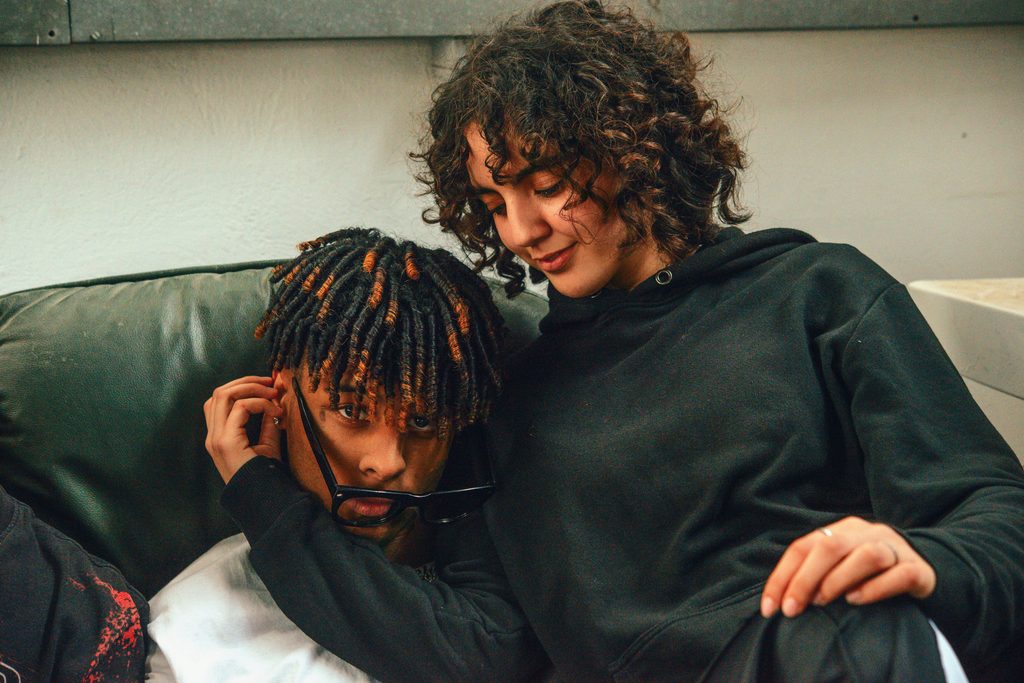A young couple sitting closely together on a couch, with the woman affectionately leaning over the man. The man has short twists with brown highlights and holds his sunglasses near his face, while both wear dark hoodies.